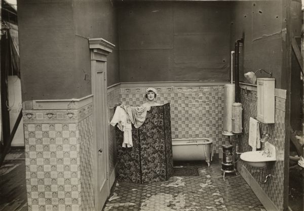 Actress Anna Luther peeps over the top of a dressing screen at the back of a bathroom set at the Keystone Studios in a production still for the 1916 short "Bath Tub Perils." The photograph shows where the scenery flats end on both sides. On the right a technician is kneeling at the back of the sink.