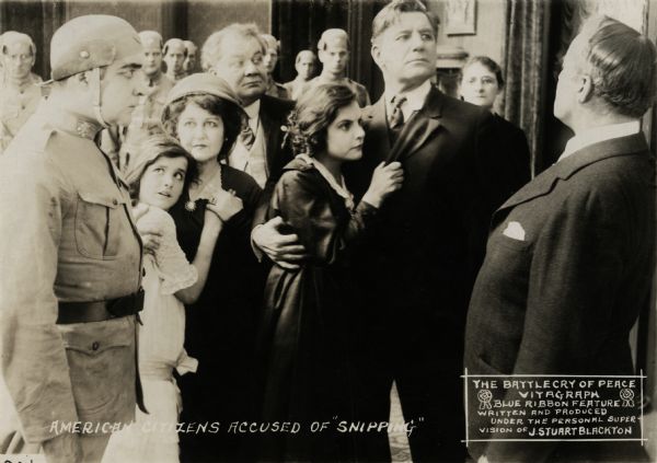 After the invasion of the United States, Americans are threatened by the enemy leader Mr. Emanon (played by L. Rodgers Lytton in profile at far right) and a room full of enemy soldiers. Closest to Mr. Emanon are John Harrison (played by Charles Richman) and Virginia Vandergriff (Norma Talmadge). Virginia's mother (played by Louise Beaudet) and little sister Dorothy (Lucille Hamill) look fearfully at the enemy soldier in the foreground (Harry Northrup). Behind Louise Beaudet is the actor James Lackaye in this scene still for the 1915 Vitagraph silent film "The Battle Cry of Peace."