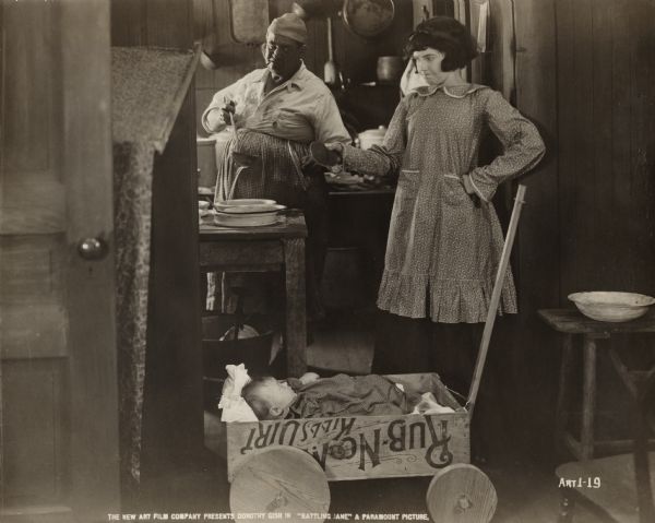 Jane the waif (played by Dorothy Gish) offers Baby Sheldon (Ernest Marion) a pancake in a scene still for the silent film "Battling Jane." Behind them is a large male cook in a kitchen.