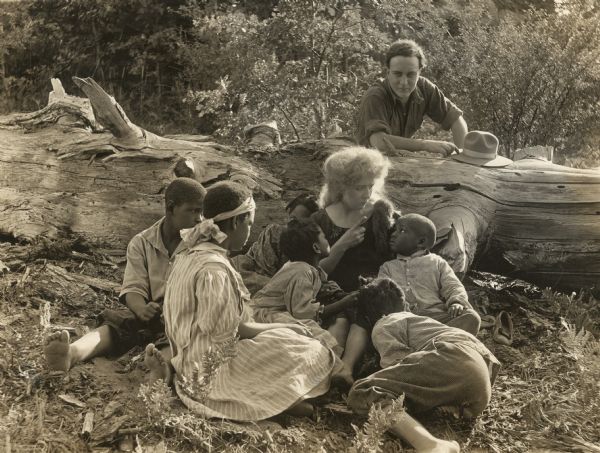 In a scene still for "Bawbs o' Blue Ridge," Barbara "Bawbs" Colby (played by Bessie Barriscale) entertains six African-American children among the trees of the Appalachian Mountains. Ralph Gunter (played by Arthur Shirley) watches from behind a fallen tree.