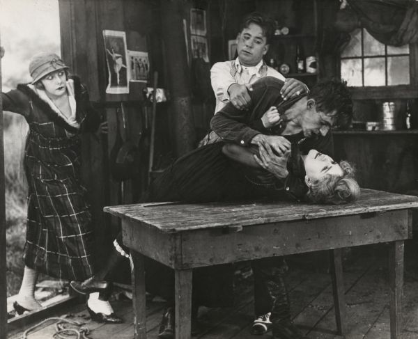 In this scene still for episode seven ("A Name for a Baby") of the silent serial "Beatrice Fairfax," Beatrice Fairfax (played by Grace Darling) bursts in upon a fight in a shack. Jimmy Barton, her colleague from the newspaper (played by Harry Fox), is grappling with the Outcast (played by Robin H. Townley) who in turn is attacking the unwed mother Madge Minturn (Mary Cranston).