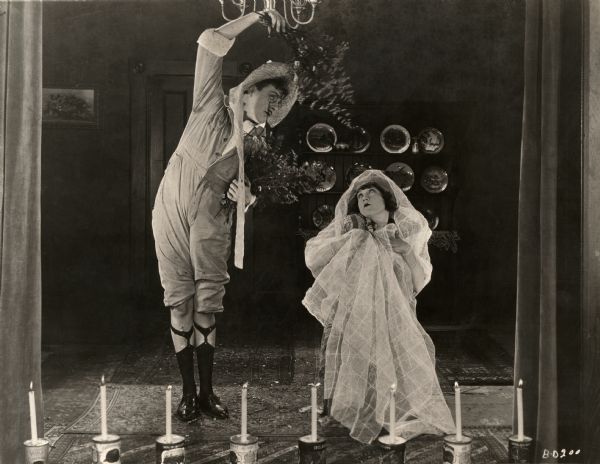 Harry Myers and Marie Prevost perform in a makeshift home theater with footlights made from candles on canned goods in a scene still for the silent drama "The Beautiful and the Damned." Myers, wearing pince nez, plays the part of a tree, while Prevost kneels and has an affectedly winsome expression on her face.