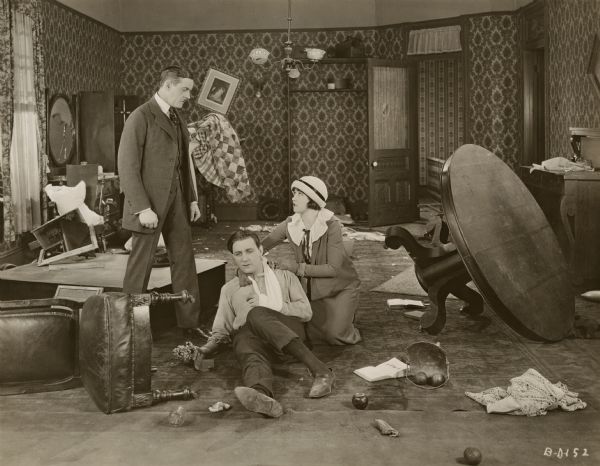 This scene still for the 1922 Warner drama "The Beautiful and the Damned" is set in a wrecked room with an overturned chair and table, broken picture frames, mirrors, and so on. On the floor are Anthony Patch, played by Kenneth Harlan with his arm in a sling, and his wife Gloria (Marie Prevost). The man belligerently standing over them  appears to be the actor Parker McConnell.
