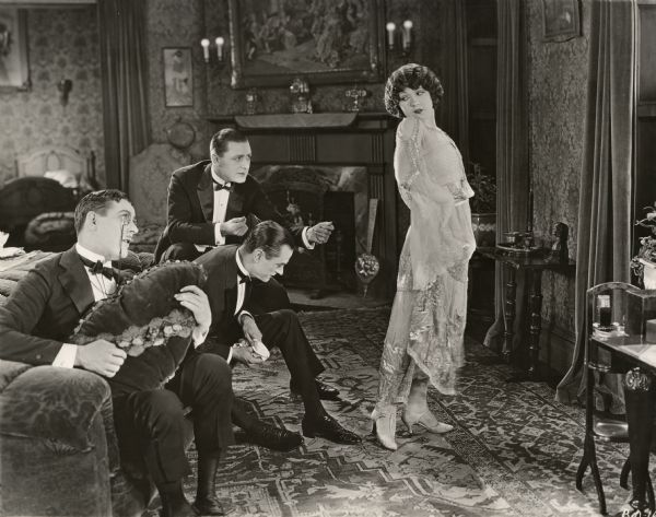 In this scene still for the silent Warner Brothers drama "The Beautiful and the Damned," three men dressed in formal evening wear show their appreciation for the legs of Marie Prevost who wears a gauzy gown with satin accents. On the left is actor Harry Myers wearing pince nez. Sitting on the arm of the sofa, snapping his fingers is Kenneth Harlan. An unidentified actor sits between them.