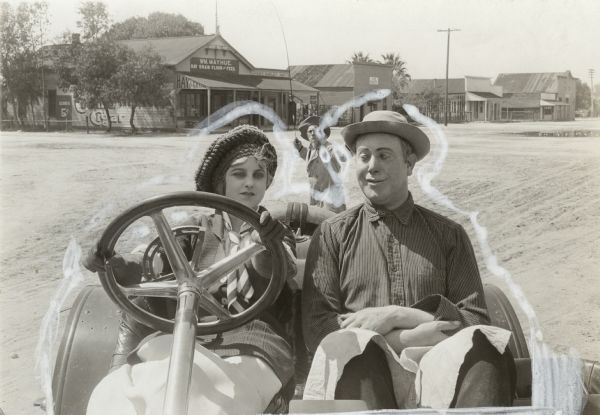 Actress Ella Hall sits behind the wheel of an open roadster automobile, perhaps a Stutz Bearcat. Sitting beside her is Robert Z. Leonard costumed for a county bumpkin role. Behind them an angry farmer brandishes a buggy whip. In the background is the William Mayhue general store in Newhall, California, in Santa Clarita north of Los Angeles.