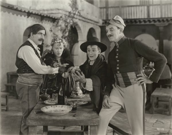 From left to right, grouped around a table in a courtyard in a scene still from the 1922 silent comedy "The Beauty Shop," are the actors James J. Corbett, Louise Fazenda, Billy B. Van, and Montagu Love.