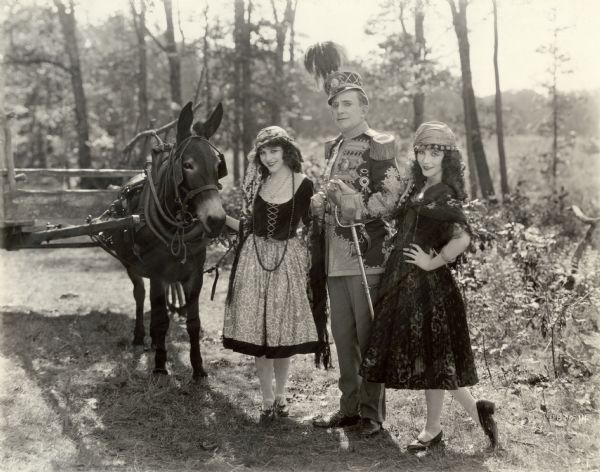 Raymond Hitchcock (in costume as Dr. Arbutus Budd pretending to be an Italian nobleman) is arm in arm with the Fairbanks Twins, Marion Fairbanks on the left and Madeline Fairbanks on the right. The twins are costumed as the Italian peasant girls Cola and Coca in this scene still for the 1922 comedy "The Beauty Shop." Marion holds the halter of a donkey harnessed to a farm cart.