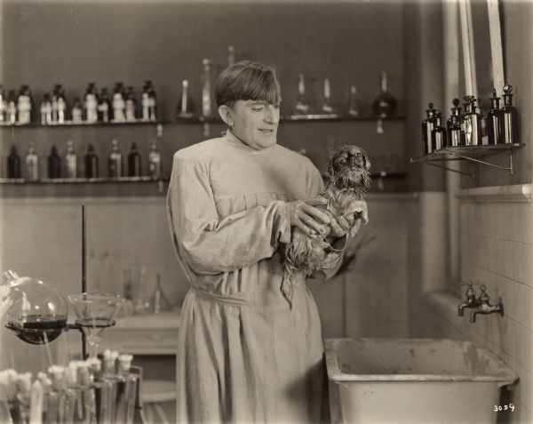 Dr. Arbutus Budd, played by Raymond Hitchcock, is in his laboratory testing a new beauty formula on a forlorn Pekingese dog in a scene still for the silent comedy "The Beauty Shop." Hitchcock is costumed in a lab coat. Around him are bottles of chemicals and lab apparatus.