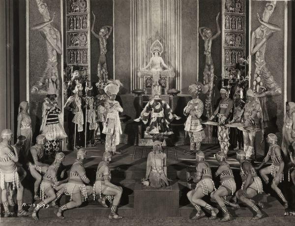 Marion Davies, costumed as an East Indian princess, sits enthroned at the back of a lavishly decorated set surrounded by a crowd of worshipful soldiers, dancing girls, servants, and nobles in a scene still for the 1922 drama "Beauty's Worth."