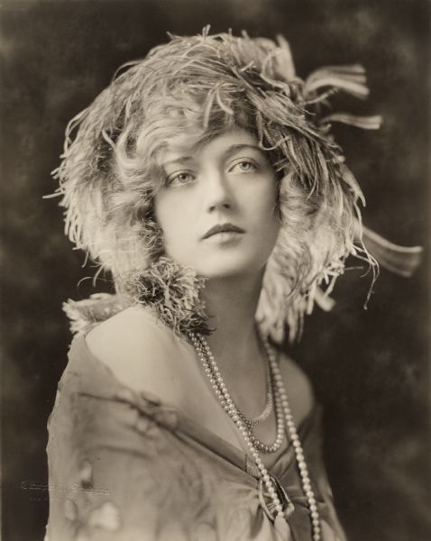 Full face, quarter-length studio portrait of the actress Marion Davies used to promote the 1922 silent drama "Beauty's Worth." She wears several strands of pearls and a headdress with feathers.