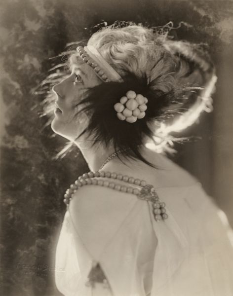 Left profile studio portrait of the actress Marion Davies used to promote the 1922 silent drama "Beauty's Worth." She wears strands of pearls and a pearl and feather headdress.