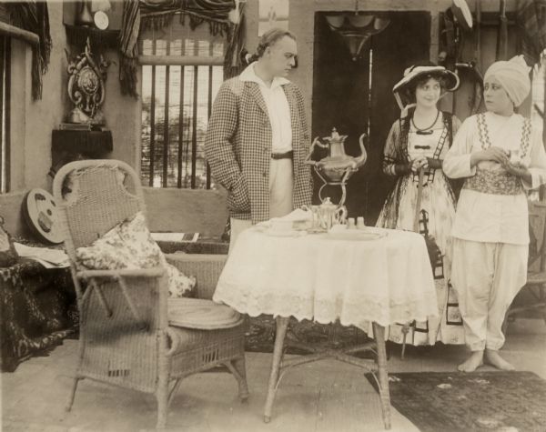 In this scene still for the silent drama "The Beckoning Flame," the Indian colonial administrator Harry Dickson (played by Henry Woodruff) and his English sweetheart Elsa Arlington (Rhea Mitchell) stand behind a tea table. They are being waited upon by Dickson's boy servant who is actually Janira, an Indian princess in disguise played by the Japanese actress Tsuru Aoki.