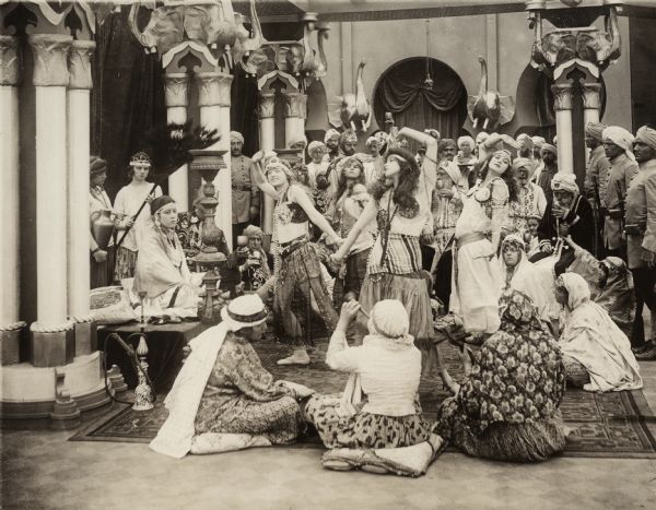 The Indian princess Janira (played by Japanese actress Tsuru Aoki) is seated on a raised platform on the left. In front of her are four dancing girls and a host of musicians, soldiers, and Indian nobles in a scene still for the 1916 silent drama "The Beckoning Flame."