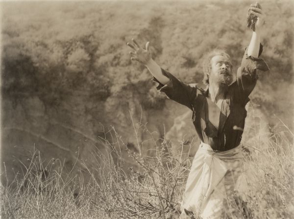 In a scene still for the silent drama "The Beggar of Crawnpore," H. B. Warner poses in character as Dr. Robert Lowndes, a British army doctor in India reduced to wild eyed beggary by morphine addiction. He stands in a field wearing a torn shirt and raises a handful of poppy straw to the sky.
