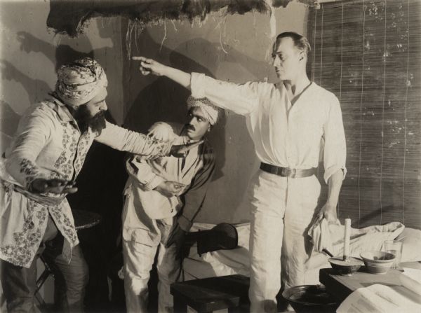 In a scene still for the silent drama "The Beggar of Crawnpore," H. B. Warner poses in character as Dr. Robert Lowndes, a British army doctor in India. He is in a bedroom and appears to be giving directions to two Indian men in turbans, apparently Sikhs.