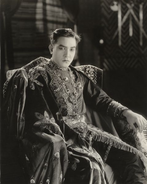 Sessue Hayakawa poses in the role of the Prince of the Island of Desire in a 3/4 length seated portrait for the 1920 silent fantasy film "The Beggar Prince." Hayakawa's costume is covered with pearls, jewels, and embroidery.