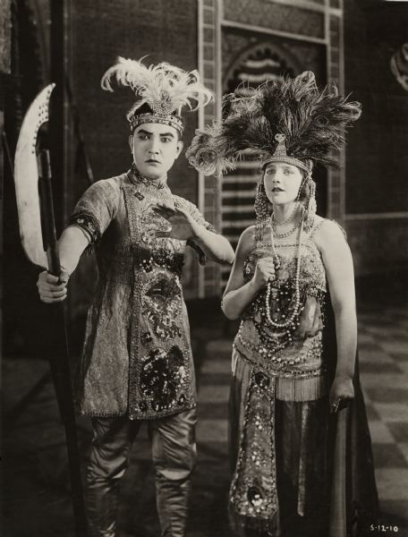 In this scene still for the silent drama "The Beggar Prince, the Prince of the Island of Desire (played by Sessue Hayakawa) and Olala (Beatrice LaPlante) wear the elaborately embroidered and bejeweled aristocratic costumes of an imaginary East Indian kingdom. Hayakawa holds the poled weapon called a bardiche.