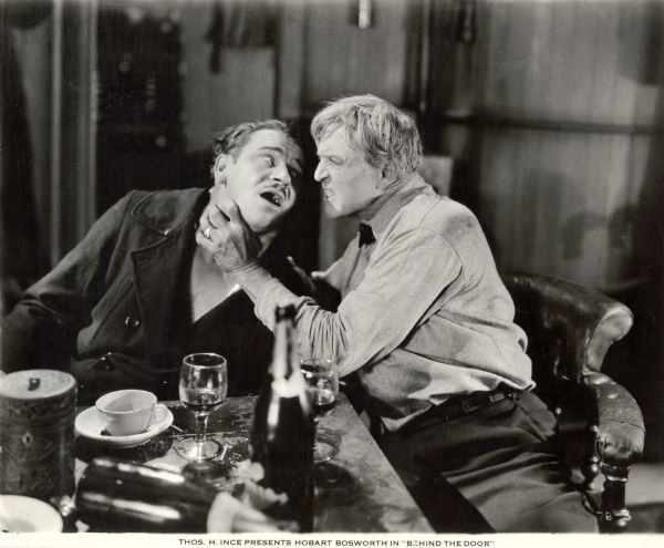 In a scene still for the silent drama "Behind the Door," German U-boat commander Lieutenant Brandt (played by Wallace Beery) is being throttled by American Merchant Marine Captain Oscar Krug (Hobart Bosworth). They are seated at a table in Krug's cabin.