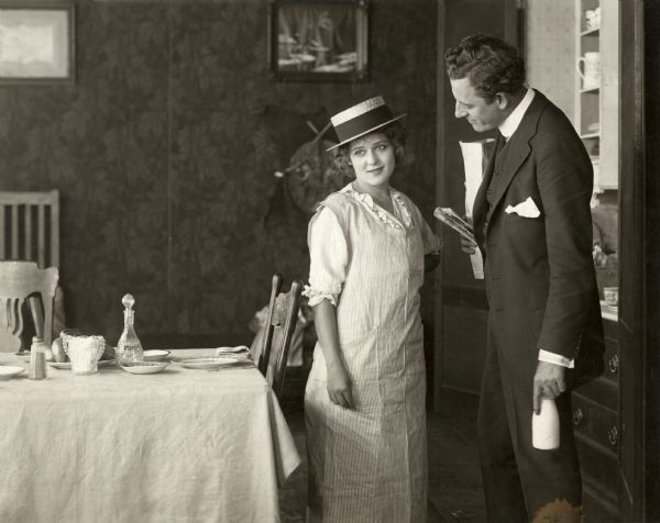 Dolly Lane (played by Mary Pickford) and her husband Steve Hunter (James Kirkwood) are in their dining room. Pickford wears an apron over her dress and a man's straw boater on her head. Kirkwood holds a newspaper and a bottle of milk.