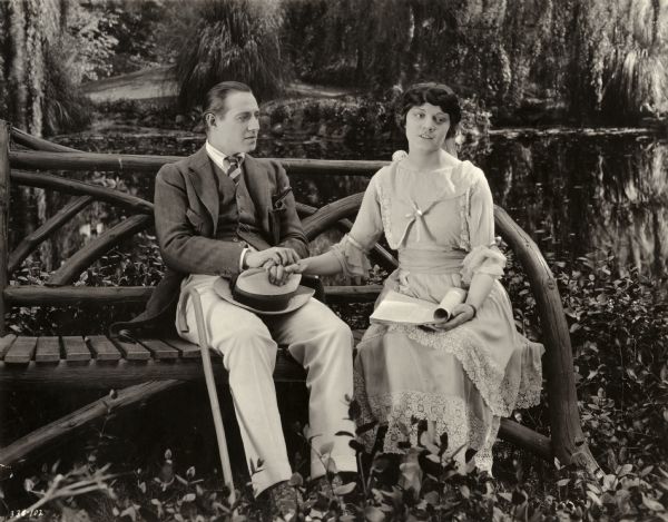 Richard Armour, played by Elliott Dexter, sits on a park bench by a pond with his brother's wife Lali Armour, played by Mabel Julienne Scott, in a scene still for the silent drama "Behold My Wife." Lali is a half-breed Indian from Canada whom Frank Armour married to humiliate his aristocratic British family, but Richard educates her much as Henry Higgins educated Eliza Doolittle in Shaw's "Pygmalion."