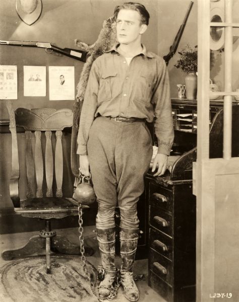 Wallace Reid, wearing jodhpurs and a work shirt, stands in a sherrif's office with a ball and chain attached to his ankle. Behind him is a rolltop desk and on the wall wanted posters and rifles.