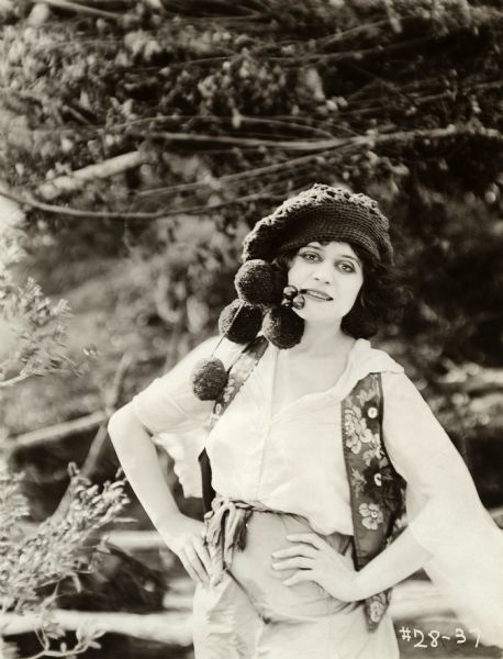 In this publicity still for the 1918 Fox silent drama "The She-Devil," Theda Bara poses outdoors with cherries hanging from her lips and four giant pom-poms hanging from her hat.