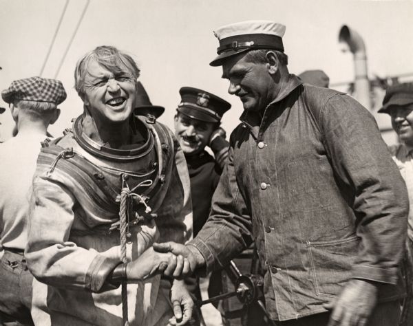 In a hardhat diving suit, actor Hobart Bosworth shakes hands with a professional diver in a publicity still for the 1920 silent drama "Below the Surface."