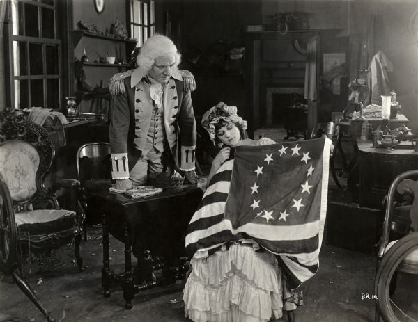 General George Washington, played by George MacQuarrie, looks on as Betsy Ross (Alice Brady) shows him the American flag she has been sewing. This is a scene still for the silent patriotic drama "Betsy Ross."