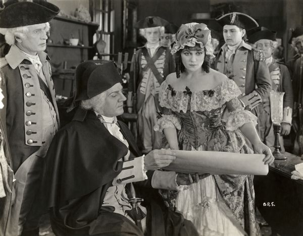 General George Washington (played by George McQuarrie) examines a document presented by Betsy Ross (Alice Brady). Over her left shoulder, in the role of Joseph Ashburn is John Bowers. Six unidentified actors dressed in American Revolutionary Army uniform are also in this scene still for the silent drama "Betsy Ross."