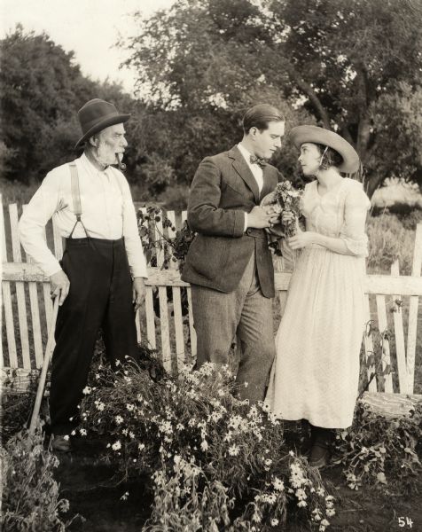 In a scruffy flower garden, Peter Van Alstyne (played by David Butler) and Nancy Scroggs (Zasu Pitts) cast loving eyes at each other while Nancy's father Ezra Scroggs (Jack McDonald) glares at them in a scene still for the 1919 silent drama "Better Times."