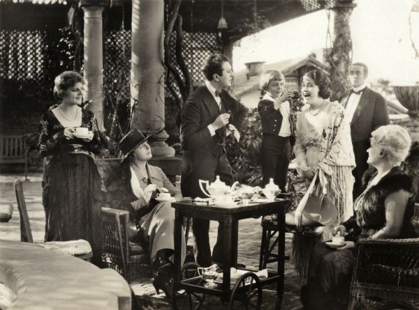 During a tea party in an English garden, Little Dick Beverly (played by the child actor Ben Alexander) feeds a piece of cake to the American visitor Charmian Page (Clara Kimball Young) in a scene still for the silent drama "The Better Wife." Around them are Lillian Walker (far left, playing Helen Kingdon), Kathlyn Williams (seated in a black hat playing Lady Beverly), and Elinor Hancock (far right). Standing with a monocle in his hand is the Comte de Cheveral, played by Irving Cummings. 	