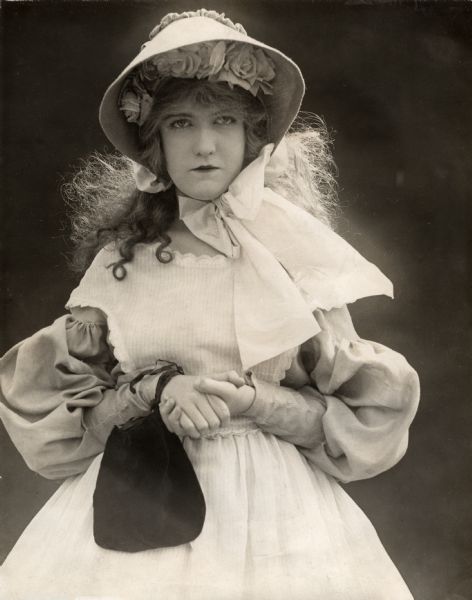 Publicity still of Dorothy Gish costumed for the role of Betty Lockwood in the silent drama "Betty of Graystone." She wears a wide bonnet with roses underneath and a pinafore over a blouse with bishop sleeves.