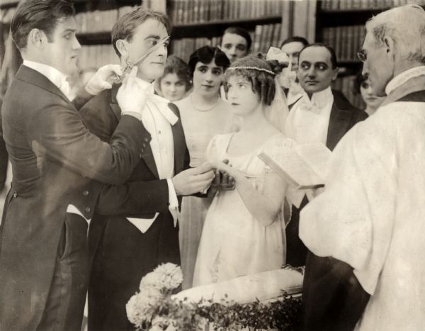 In this scene still for the silent drama "Betty of Graystone," David Lockwood (played by Owen Moore) weds Betty (Dorothy Gish) in a home ceremony with a priest and witnesses. At far left is the actor Warner Richmond who, curiously, holds a knife against Moore's cheek. Between Moore and Gish is the actress Leonore Harris. The other players are unidentified.