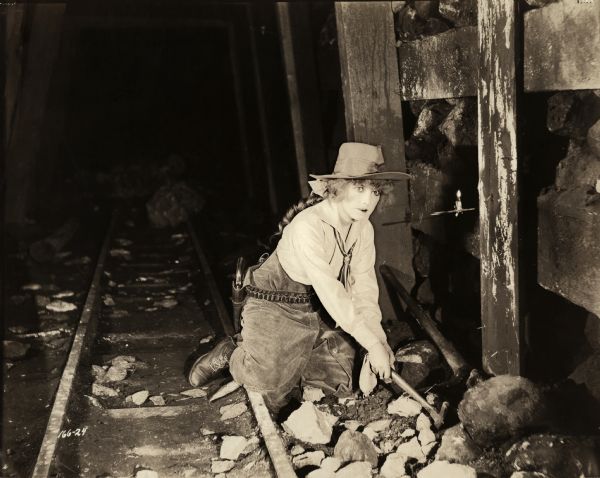 Betty Sherwin, played by Fannie Ward with her rock pick in hand, searches her inherited mine for gold in a scene still for the silent comedy "Betty to the Rescue." She is in western wear: cowboy hat, corduroy riding breeches, and a cartridge belt with a revolver.