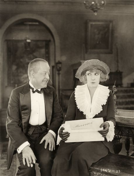 The banker J. Peter Weathersby (played by Louis Haines grinning with a cigar in his hand) sits beside Sally Marrio (Pearl White) who is delivering a pair of shoes from her husband's firm in a scene still from the silent melodrama "Beyond Price."