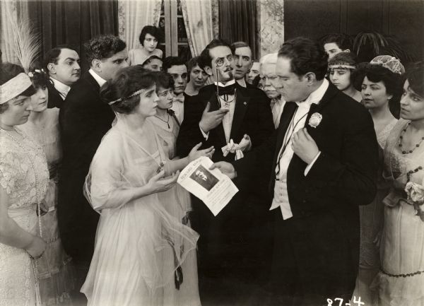 Surrounded by a crowd of extras in evening clothes, a police officer (played by Arthur Bauer) holds out a wanted poster reading "Reward $50,000 Charles Burliardi" to Bianca Wells, played by Florence La Badie. Count Berdeau, played by Charles A. Jahn wearing a monocle, protests that he is not the criminal being sought in this scene still for the silent drama "Bianca Forgets."