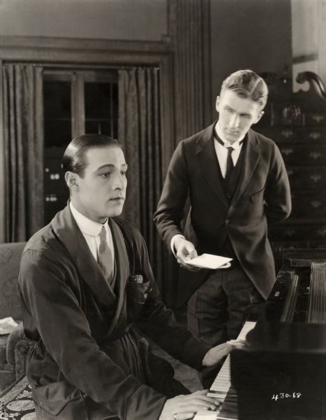 Lord Hector Bracondale (played by Rudolph Valentino) sits in a reverie at a piano as a butler delivers a letter on a salver in a scene still for the silent drama "Beyond the Rocks."