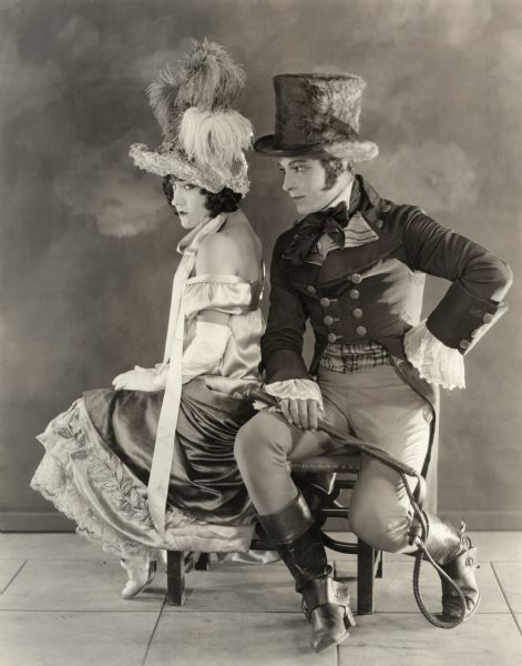 Gloria Swanson, as Theodora Fitzgerald, and Rudolph Valentino, as Lord Hector Bracondale, in a full-length seated Keyes studio publicity still for the 1922 silent drama "Beyond the Rocks." Swanson's feathery hat and Valentino's furry top hat are remarkable. He clutches a whip and she seems a bit concerned.