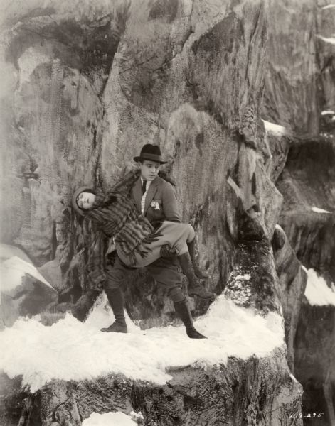Gloria Swanson (as Theodora Fitzgerald) is carried in the arms of Rudolph Valentino (Lord Hector Bracondale) in her rescue from a snowy mountain ledge in a scene still for the 1922 silent drama "Beyond the Rocks." They are costumed in aristocratic outdoor gear. Valentino wears a homburg hat, jodhpurs, and a checked waistcoat. Swanson is in a short mink coat and wool skirt. Both wear high boots.