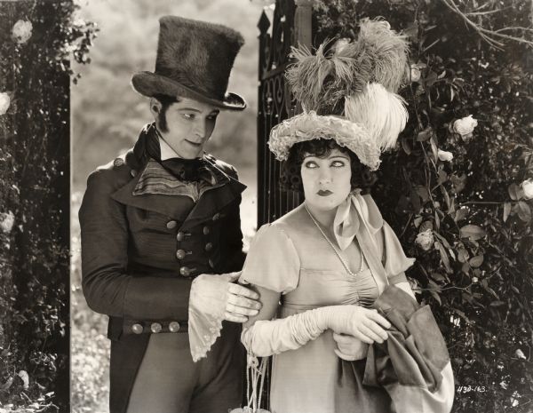Lord Hector Bracondale, played by Rudolph Valentino, approaches Theodora Fitzgerald (Gloria Swanson) by a gate in a garden wall covered with camellias. They wear 18th century costumes, most notably Swanson's feathery hat and Valentino's furry top hat, in this scene still for the silent drama "Beyond the Rocks."