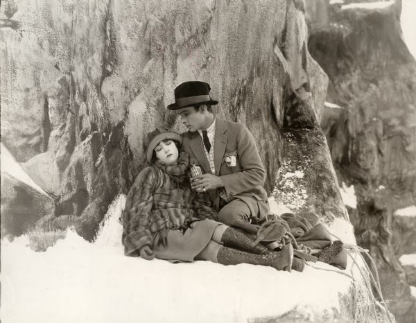 Gloria Swanson (as a semiconscious Theodora Fitzgerald) is offered a sip from the silver hip flask of Rudolph Valentino (Lord Hector Bracondale) in her rescue from a snowy mountain ledge in a scene still for the 1922 silent drama "Beyond the Rocks." They are costumed in aristocratic outdoor gear. Valentino wears a homburg hat, jodhpurs, and a checked waistcoat. Swanson is in a short mink coat, wool skirt, and high boots.