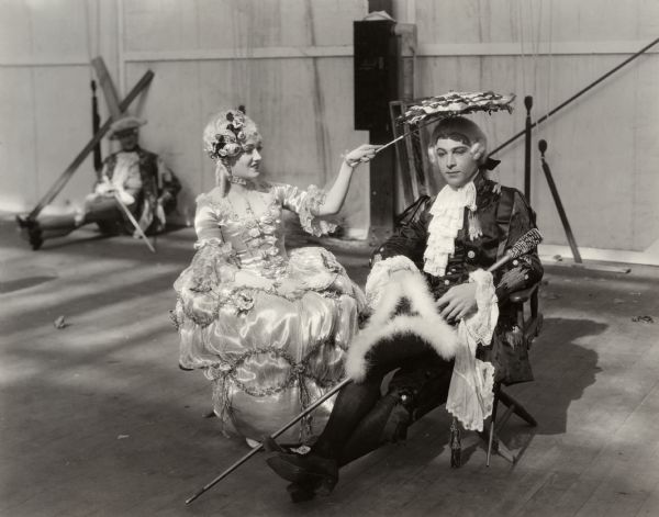 In this production still for the silent drama "Beyond the Rocks," Gloria Swanson costumed in an elaborate 18th century satin dress shades the head of Rudolf Valentino with her parasol. They are sitting in folding directors' chairs resting between scenes.