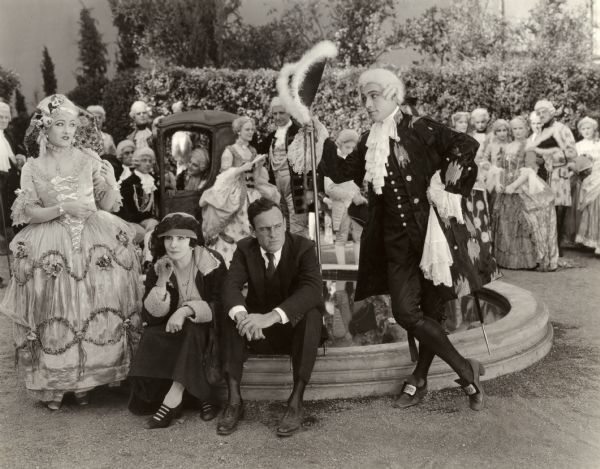 Costumed for an 18th century fantasy scene for the silent drama "Beyond the Rocks" are Gloria Swanson, Rudolf Valentino, and a host of extras in the background. Between them, seated on the edge of a fountain are Elinor Glyn who wrote the novel the film was based upon and Sam Wood the director.