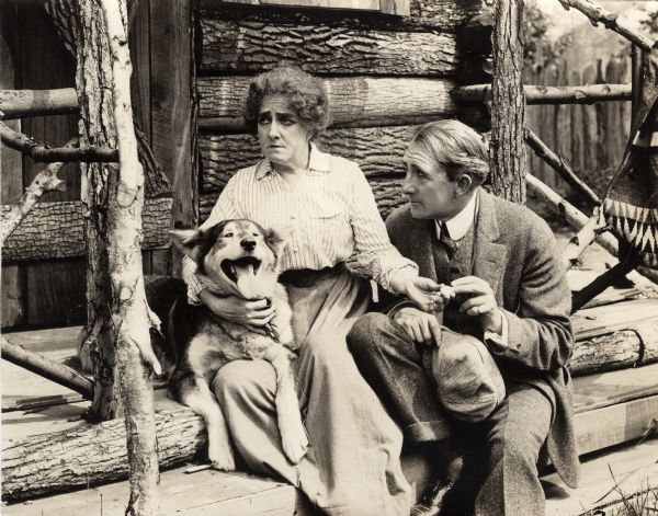 On a rustic front porch sit the actors Julia Stuart and Alec B. Francis along with a husky dog in a scene still for the silent short drama "Big Hearted Jim."