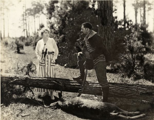 In the forest, Stella Benton (played by Kathlyn Williams in a striped skirt and white blouse) talks to Jack Fyfe (played by Wallace Reid in outdoors clothes) in a scene still for the silent drama "Big Timber." Reid has a Winchester repeating rifle. At his feet is a dead deer.