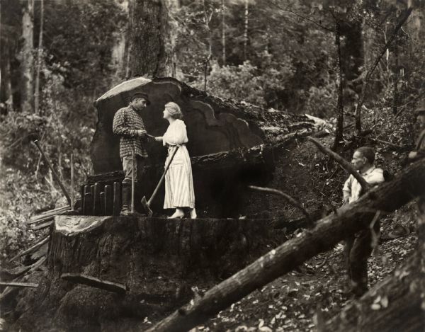 In the depths of the forest, the lumberman Jack Fyfe (played by Wallace Reid) and his wife Stella Barton (Kathlyn Williams) stand on a huge tree stump in a scene still for the silent drama "Big Timber." Reid wears a boldly checked jacket with a bib front. At their feet are lumbermen's tools: two-bitted axe, mauls, wedges.