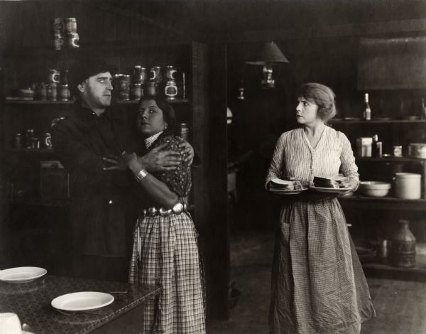 Lumberman Jack Fyfe (played by Wallace Reid) is caught embracing Neemis, the Indian kitchen maid, by his wife Stella Barton (Kathlyn Williams) in the cookhouse of his lumber camp in a scene still for the silent drama "Big Timber."