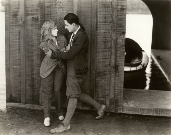 Grayson Blair (played by Alan Forrest) discovers that the boy he has seized is actually Phyllis King (played by Mary Miles Minter) dressed in her brother's clothes. This scene still for the silent drama "A Bit of Jade" is set on the waterfront near a boat house. Minter wears a checked jacket. Forrest wears knickerbockers and a cardigan with shawl collar.