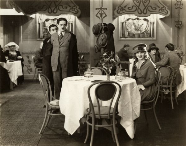 As Grayson Blair (played by Alan Forrest) is helped with his coat, he notices Phyllis King (Mary Miles Minter) sitting alone at a table in a restaurant in this scene still for the silent drama "A Bit of Jade."