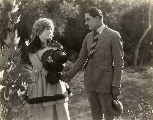 Wearing a fur-trimmed coat, Phyllis King (played by Mary Miles Minter) is shaking hands with Grayson Blair (Alan Forrest) in a scene still for the silent drama "A Bit of Jade." Forrest is wearing a tweed Norfolk suit, striped necktie, and cloth cap.